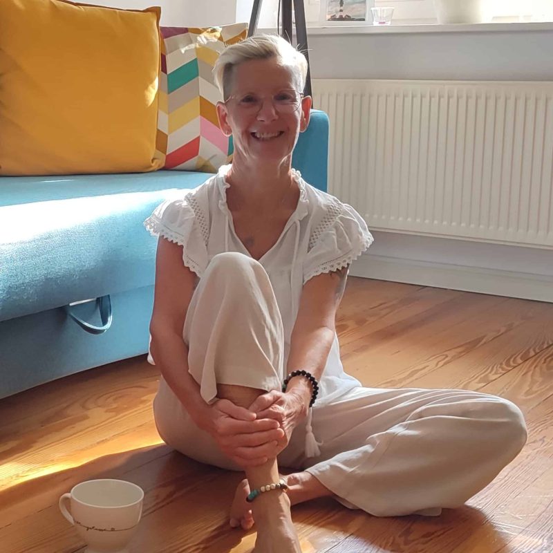 Daniela Bernauer sitzt barfuß entspannt mit einer Tasse Kaffee lachend auf dem Holzboden – Elterncoach offen, herzlich und natürlich.