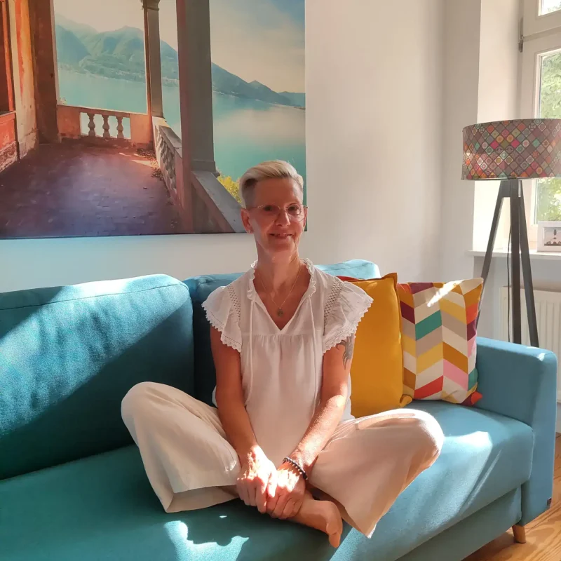 Die Elternbegleiterin von KinderLiebe sitzt entspannt im sonnendurchfluteten Wohnzimmer auf einem türkisfarbenen Sofa – ein warmer Ort, an dem Eltern sich verstanden fühlen können.