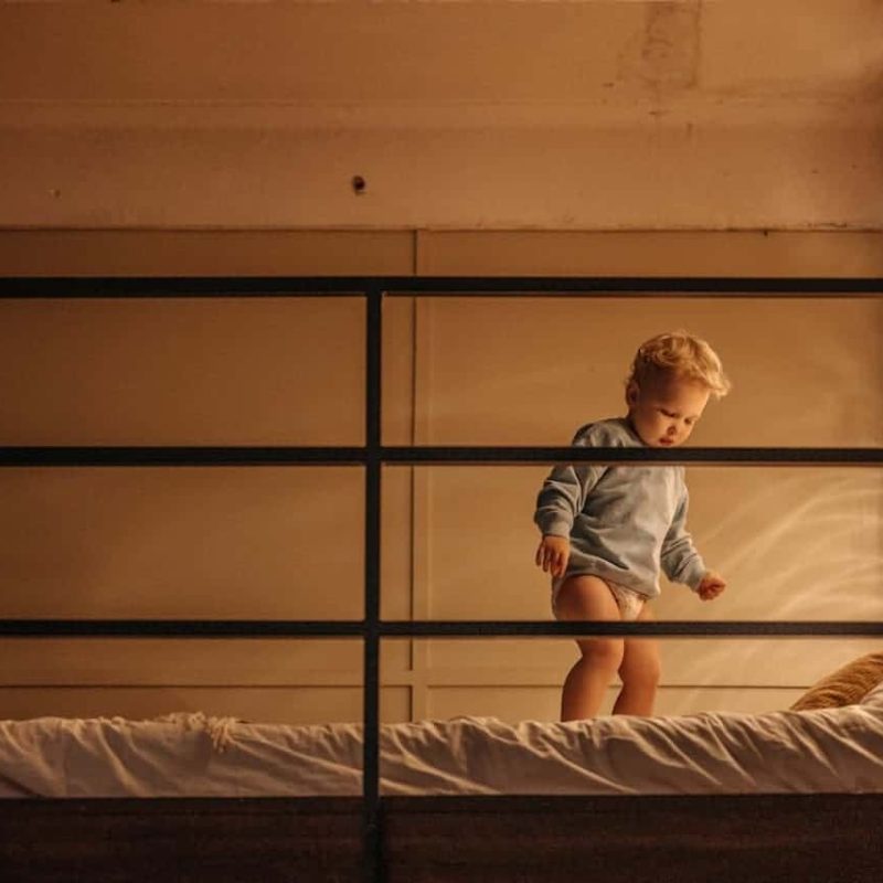 Kleinkind steht in Windel auf Bett bei warmem Licht – Alltag vor dem Trockenwerden.