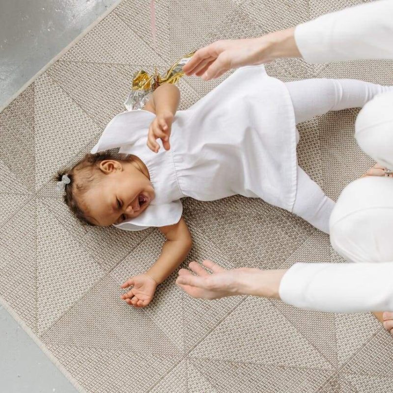 Kleinkind liegt wütend auf dem Boden und schreit während eine erwachsene Hand beruhigend nach ihm greift.